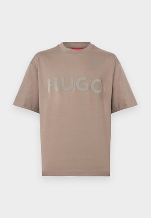 Beige lyhythihainen t-paita pyöreällä kaula-aukolla ja suurella kohopainetulla "HUGO"-tekstillä rinnassa yksivärisellä taustalla.