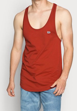Man draagt een mouwloos rood tanktop met een klein "Lee"-logo op de borst en lichtblauwe spijkerbroek, staand tegen een effen achtergrond.