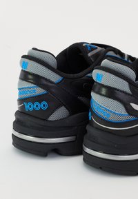 Paire de baskets New Balance 1000 noires et grises avec des accents bleus, logos visibles et semelles épaisses et rembourrées, vues de l'arrière.