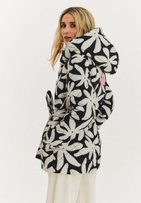 Černá prošívaná bunda s velkým bílým květinovým vzorem, s kapucí, páskem v pase a jemnou texturou. Pohled zezadu zdůrazňuje detaily designu.