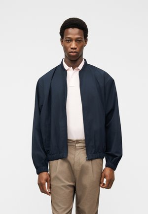 Hombre vestido con una chaqueta azul marino con cremallera sobre una camisa tipo polo rosa claro y pantalones beige, de pie contra un fondo claro y liso.