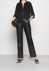 Schwarze durchsichtige Bluse mit tiefem V-Ausschnitt und gerafftem Kragen; schwarze Leder, gerade geschnittene Hose; schwarze Sandalen mit offenen Zehen und Knoten-Details; schwarze Clutch.