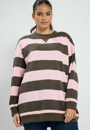 Femme aux cheveux foncés attachés en queue de cheval, portant un pull oversize avec de larges rayures horizontales roses et marron, associé à un pantalon foncé.