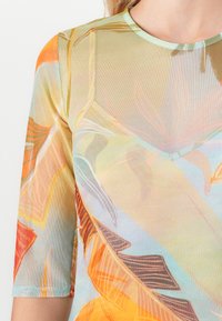 Close-up van een doorschijnende top met een meerkleurig tropisch bladpatroon in oranje, geel, blauw en bruin op een lichte achtergrond.