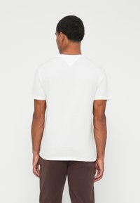 Hombre con cabello corto que lleva una camiseta blanca de manga corta sencilla y pantalones marrón oscuro, de pie de espaldas contra un fondo blanco.