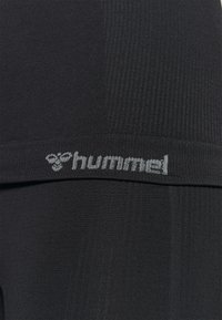 Close-up of black ribbed fabric with grey "hummel" logo and bee symbol on the waistband area of clothing.