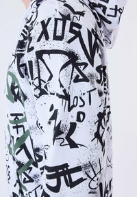 Sudadera blanca con una mezcla de texto y símbolos en estilo graffiti en negro y verde. Tejido suave con patrones abstractos en toda la superficie.