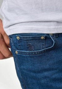 Kék farmernadrág, amelyen az elülső zseb kis hímzett logóval díszített, aranyszínű fém gombokkal és sima textúrával.