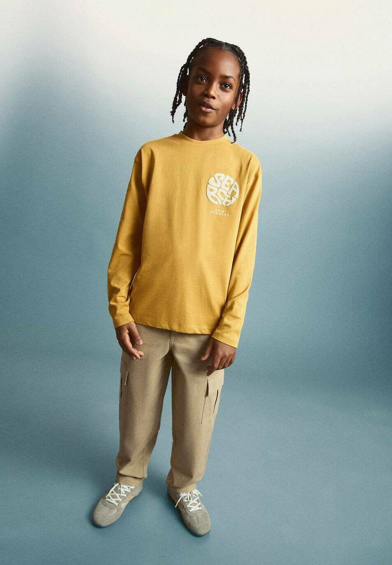 Garçon aux cheveux tressés se tient devant un fond bleu-gris, portant un chemise jaune à manches longues, un pantalon cargo beige et des baskets grises.