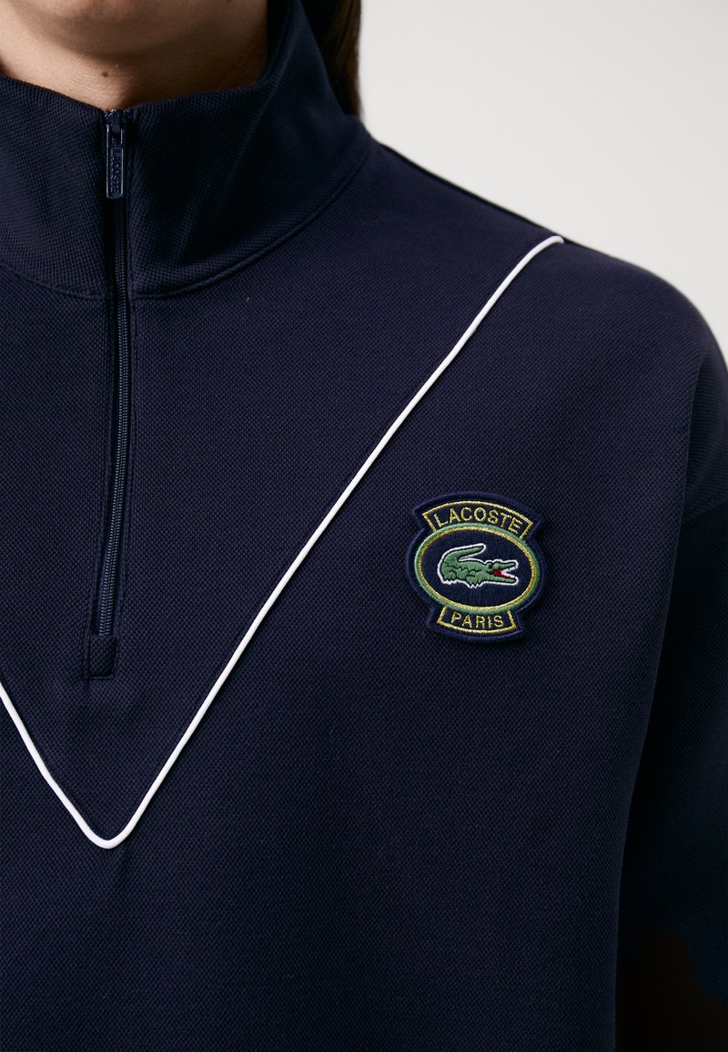 Polo marine avec col haut, fermeture à mi-zip, passepoil blanc et logo Lacoste brodé en vert représentant un crocodile et "Paris". Tissu texturé.