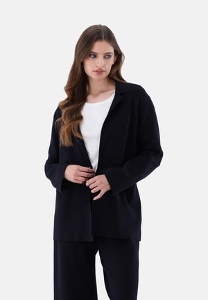 Navyblaues, oversized Blazer mit Schalkragen, langen Ärmeln und Knopfverschluss, kombiniert mit einem weißen Hemd. Glatte Textur, minimalistisches Design.