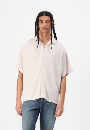 Light beige short-sleeve button-up shirt with a relaxed fit. Features a collar and subtle texture, paired with blue jeans.