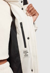 Doudoune avec un extérieur crème, arborant une doublure noire, une poche zippée et une étiquette indiquant "Un câlin chaud fait de tissu."