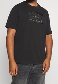 Camiseta negra de manga corta con un parche rectangular de logo que lleva el texto "TOMMY HILFIGER" y un emblema multicolor en la parte delantera.