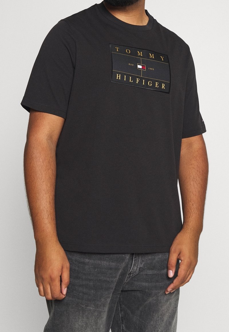 Camiseta negra de manga corta con un parche rectangular de logo que lleva el texto "TOMMY HILFIGER" y un emblema multicolor en la parte delantera.