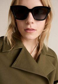 Black oversized rectangular sunglasses with gradient lenses, worn with an olive green coat featuring a notched collar and visible stitching details.