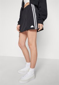 Crna suknja s bijelim Adidas trakama sa strane, izrađena od laganog materijala. Ima podesive vezice na rubu.