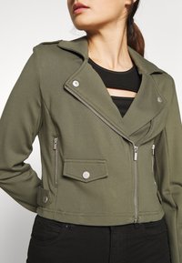 Giacca corta verde oliva con zip frontale, colletto a risvolto, hardware argentato e tasche frontali; presenta un design aderente con una texture liscia.