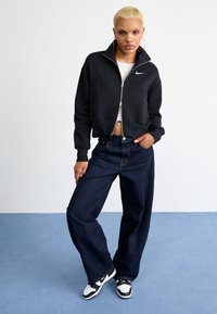 Sudadera negra con cremallera, diseño corto; jeans de mezclilla azul oscuro de talle alto; zapatillas negras y blancas; modelo posa con una mano en la cadera.