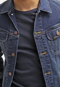 Jeansowa kurtka Lee z brązowymi guzikami noszona na czarnej koszulce z okrągłym dekoltem, eksponująca pierś i górną część ciała.