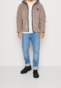 Giacca di pile marrone con zip nere, camicia bianca con colletto, jeans azzurri chiari e stivali neri stringati. Design semplice con logo ricamato.