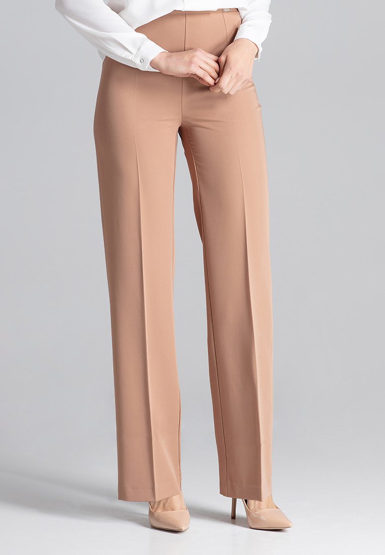 Vrouw in beige wijde broek, beige hoge hakken en een wit longsleeve shirt, staand tegen een grijze achtergrond.