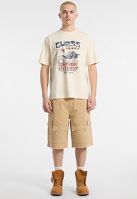 T-shirt grafico crema da uomo con testo blu e rosso, abbinato a bermuda beige con molte tasche e stivali marroni con lacci.