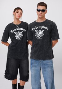 To personer har på seg matchende svarte t-skjorter med grafisk trykk i en falmet, slitt stil. T-skjortene har hvit tekst og et engeldesign. Den ene har på seg svarte denimshorts; den andre har lyseblå jeans.