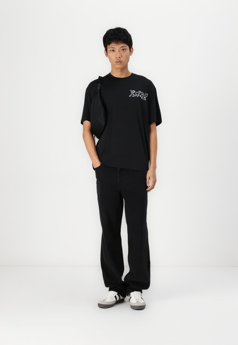 Camiseta de algodón negra con estampado gráfico en el pecho, combinada con pantalones negros holgados y zapatillas gris y blancas. El modelo lleva un bolso negro.