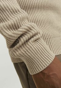 Maglione lavorato a maglia in beige chiaro con texture a coste, caratterizzato da maniche con polsini e distintivi motivi verticali. Aspetto morbido e caldo.