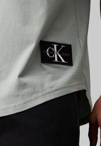 Světle šedý lem košile s černou nášivkou loga Calvin Klein Jeans nad černými kalhotami a viditelnou hnědou rukou na boku.
