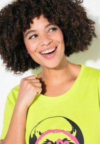 T-shirt vert néon avec un col rond et un design graphique noir/rose. Le modèle affiche des cheveux bouclés et une expression confiante.