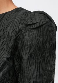 Pogled od blizu na gornji del rame osebe, ki nosi črn zgornji del iz teksturiranega blaga s povešeno, napihnjeno rokavno podrobnostjo na enobarnem ozadju.