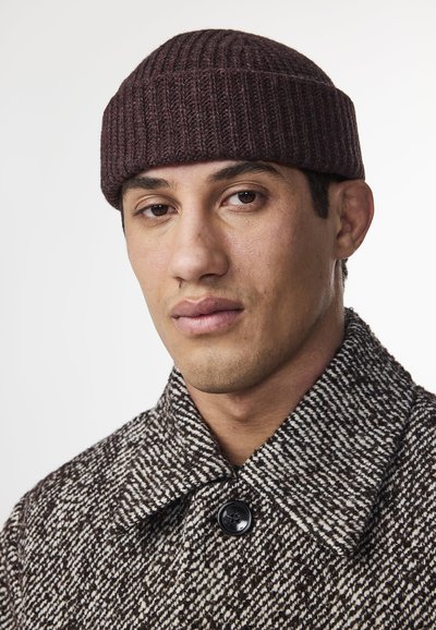 Brun og beige mønstret uldfrakke med en sort knap, parret med en maroon ribbet strikhue. Nærbillede, der viser ansigtstræk.