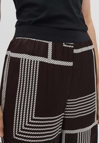 Bruine geblokte broek met witte geometrische strepen, voorzien van een zwarte elastische tailleband. Glad, lichtgewicht materiaal met een relaxte pasvorm.