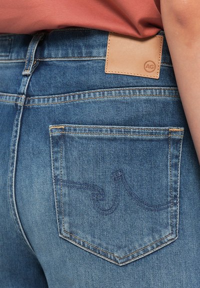 Close-up do bolso traseiro de um par de jeans de algodão azuis, com um patch de couro rotulado "AG" e um design em forma de onda bordado no bolso.