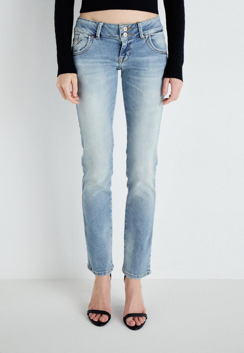 Femme portant un jean slim bleu clair avec fermeture à double bouton, associé à des talons ouverts noirs et un haut court noir à manches longues.