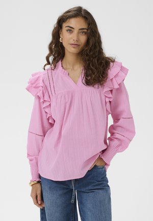 Kobieta z długimi kręconymi włosami, ubrana w różową bluzkę z falbankami i marszczonymi mankietami oraz niebieskie dżinsy, stoi z jedną ręką w kieszeni.