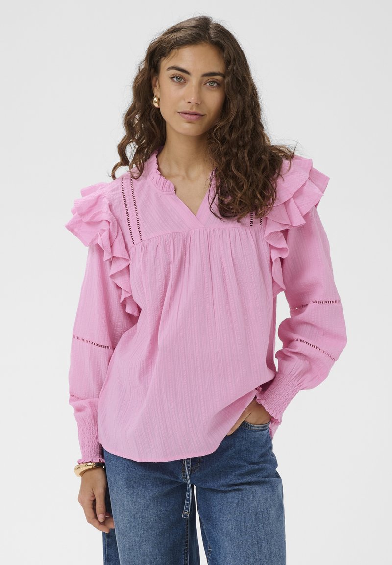 Femme aux longs cheveux bouclés portant un chemisier rose à volants avec des poignets froncés et un jean bleu, debout avec une main dans la poche.