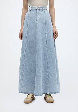 Donna che indossa una gonna lunga in denim azzurro chiaro a vita alta abbinata a sneakers beige e un top beige senza maniche, in piedi su uno sfondo semplice.