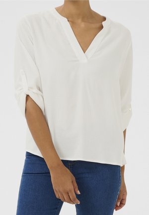 Frau trägt weiße, strukturierte V-Ausschnitt-Bluse mit hochgekrempelten 3/4-Ärmeln und blaue Jeans, steht vor einem einfarbigen Hintergrund.