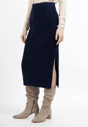 Frau trägt einen marineblauen gestrickten Midirock mit seitlichem Schlitz und beige wildlederne kniehohe Stiefel mit Absatz auf weißem Hintergrund.