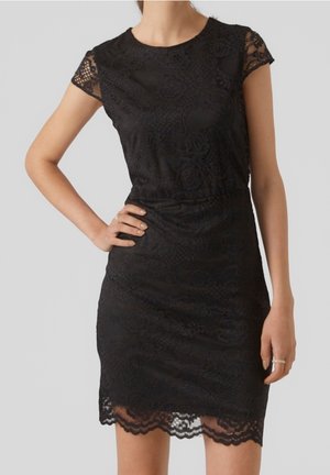 Femme portant une robe noire en dentelle ajustée avec des manches courtes et un ourlet festonné, debout avec une main sur la hanche devant un fond uni.