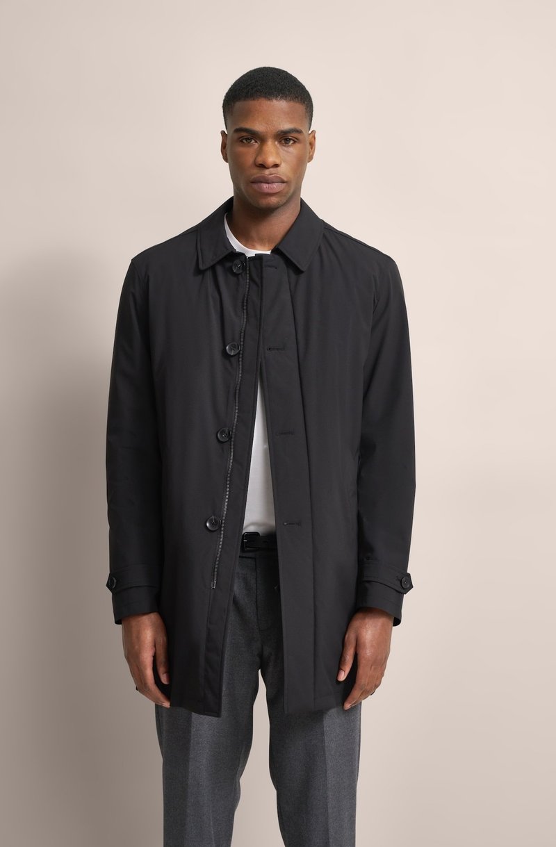 Schwarzer Trenchcoat mit Knopfverschluss, Reverskragen und Seitentaschen. Trägt man über einem weißen Hemd und grauen Hosen. Glatte Stoffoberfläche.