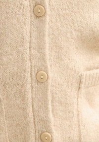 Cárdigan de punto en suave tela beige con acabado texturizado, que presenta cinco botones en tono beige claro en la parte delantera y un bolsillo en el lateral.
