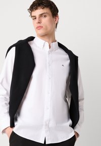 Λευκό πουκάμισο με γιακά, διακοσμημένο με κεντημένο λογότυπο. Μαύρος πλεχτός σκούφος ριγμένος στους ώμους. Απλά μαύρα παντελόνια.