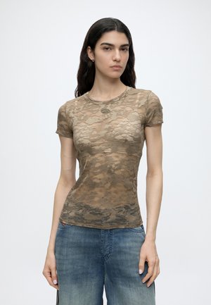 Donna con lunghi capelli scuri indossa un top trasparente in pizzo marrone a maniche corte e jeans blu, in piedi contro uno sfondo chiaro e semplice.
