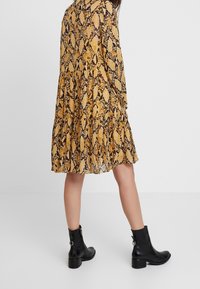 Šaty se žluto-černým hadím potiskem, s vrstveným designem, dlouhými rukávy a volným střihem. Černé kotníkové boty s ribkovanou texturou.