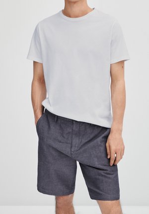 Homme portant un t-shirt blanc uni à manches courtes et un short gris foncé jusqu'aux genoux, mains dans les poches, debout devant un fond clair.