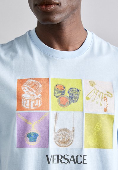 Ljusblå t-shirt med ett rutnät av färgglada smyckesillustrationer. Nyckelfärger inkluderar orange, gult, lila och grönt. "Versace" tryckt i botten.